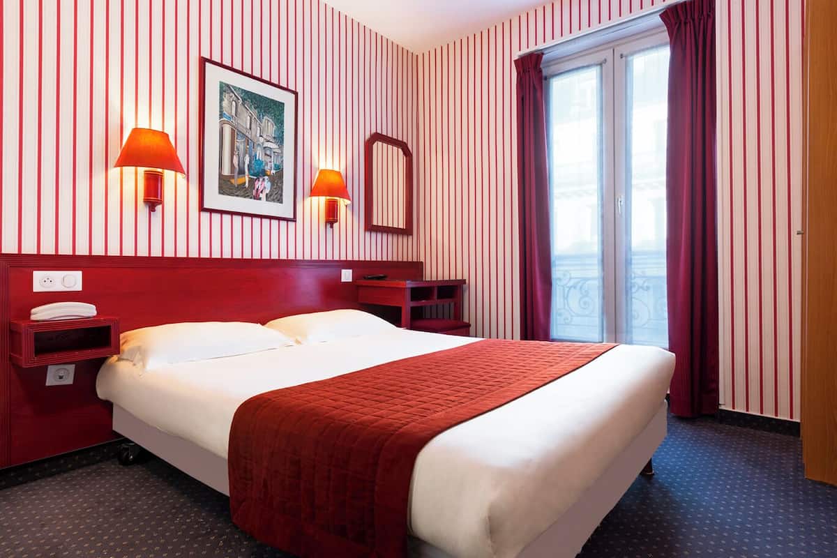 A hotel room with a large bed, two bedside tables, a nightstand, a window with curtains, and a painting on the wall.