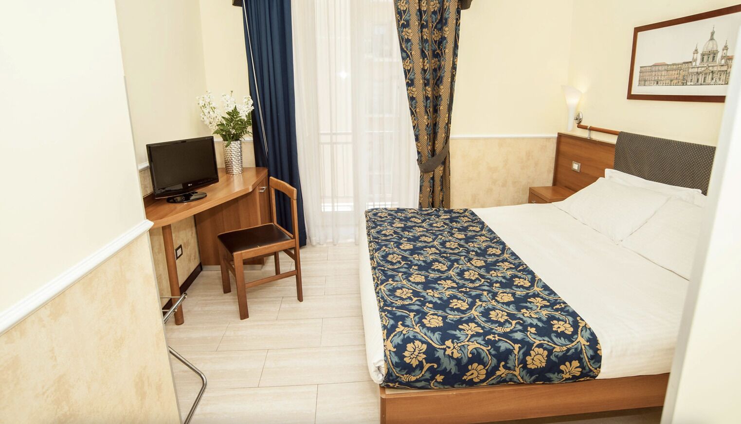 A hotel room with a bed, a desk with a computer, a chair, and a nightstand with a lamp.