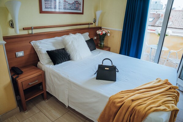 Une chambre d’hôtel avec un lit, une table de chevet, un téléphone, un vase de fleurs et une fenêtre donnant sur des immeubles.