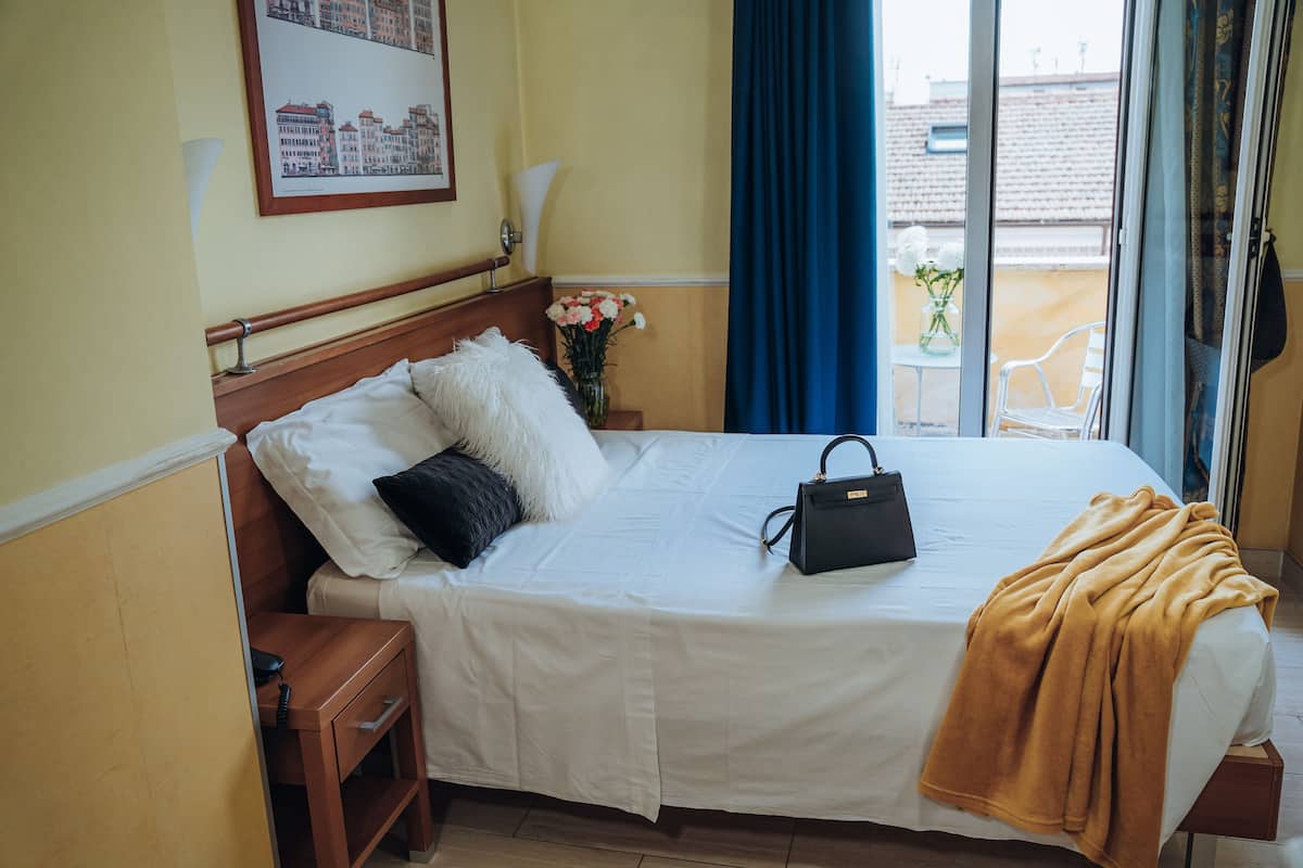 Une chambre d’hôtel avec un lit, une table de chevet, une lampe, une fenêtre avec des rideaux bleus et une vue sur les bâtiments environnants.