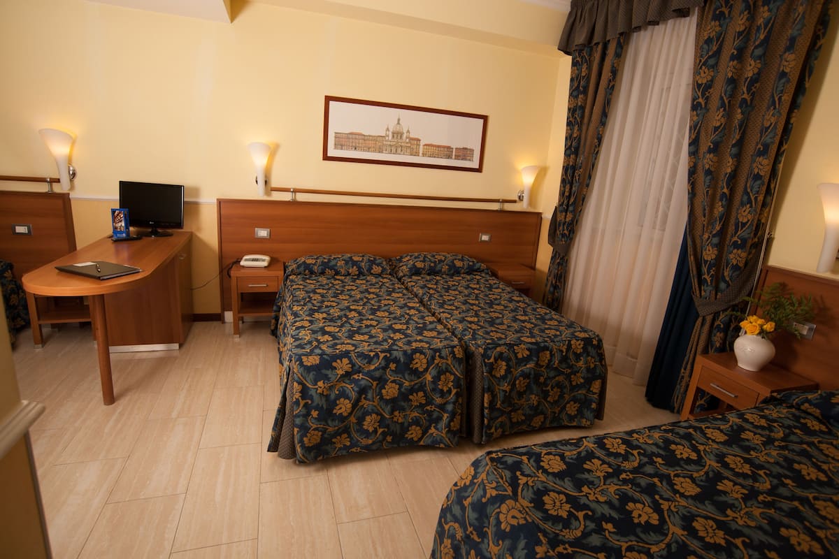 Une chambre d’hôtel avec un bureau en bois, un lit recouvert d’une literie à motifs floraux, une table de chevet avec une lampe et un tableau encadré au mur.
