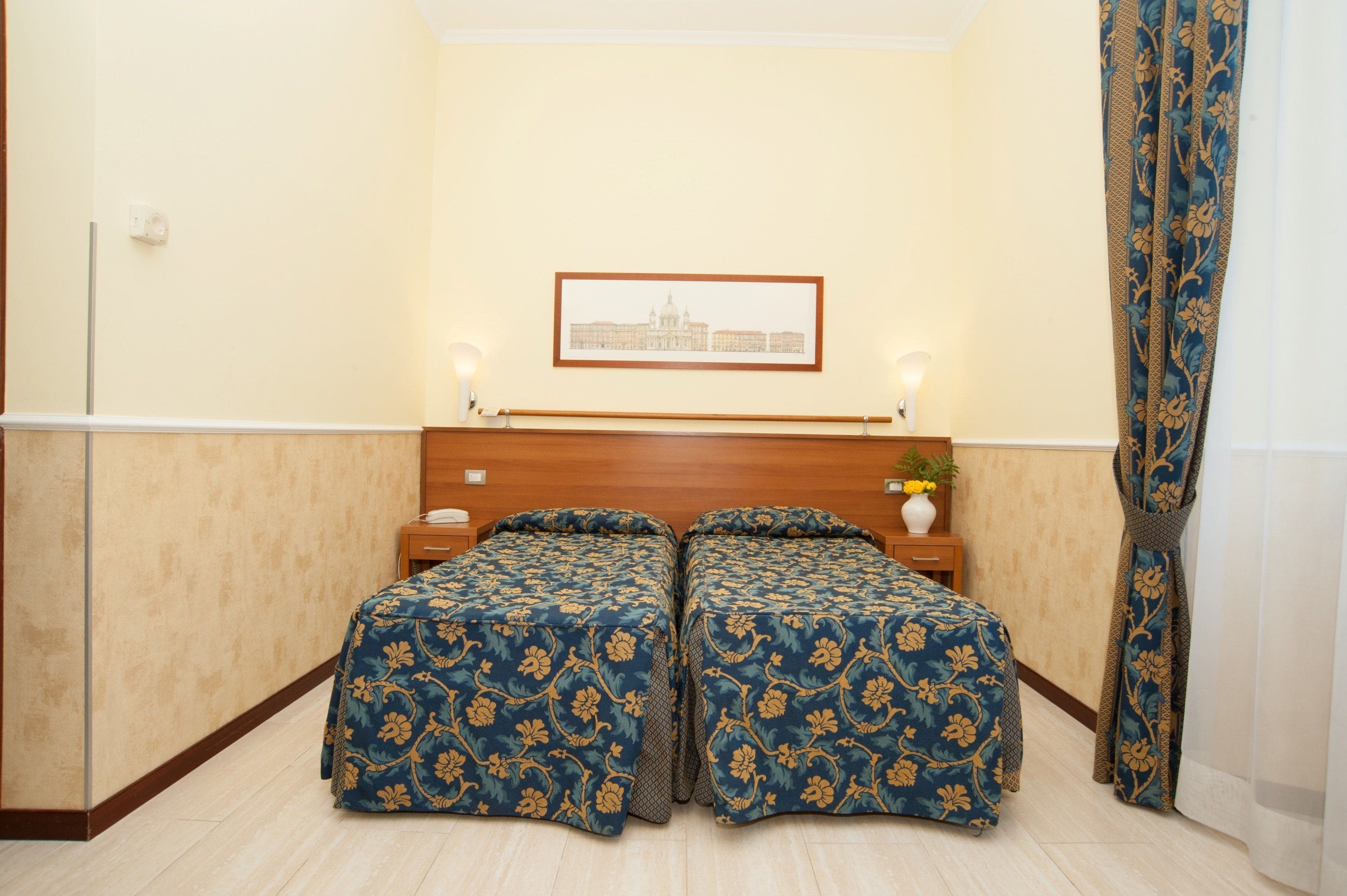 A hotel room with a double bed, a nightstand with a lamp, a framed picture on the wall, and a curtain.