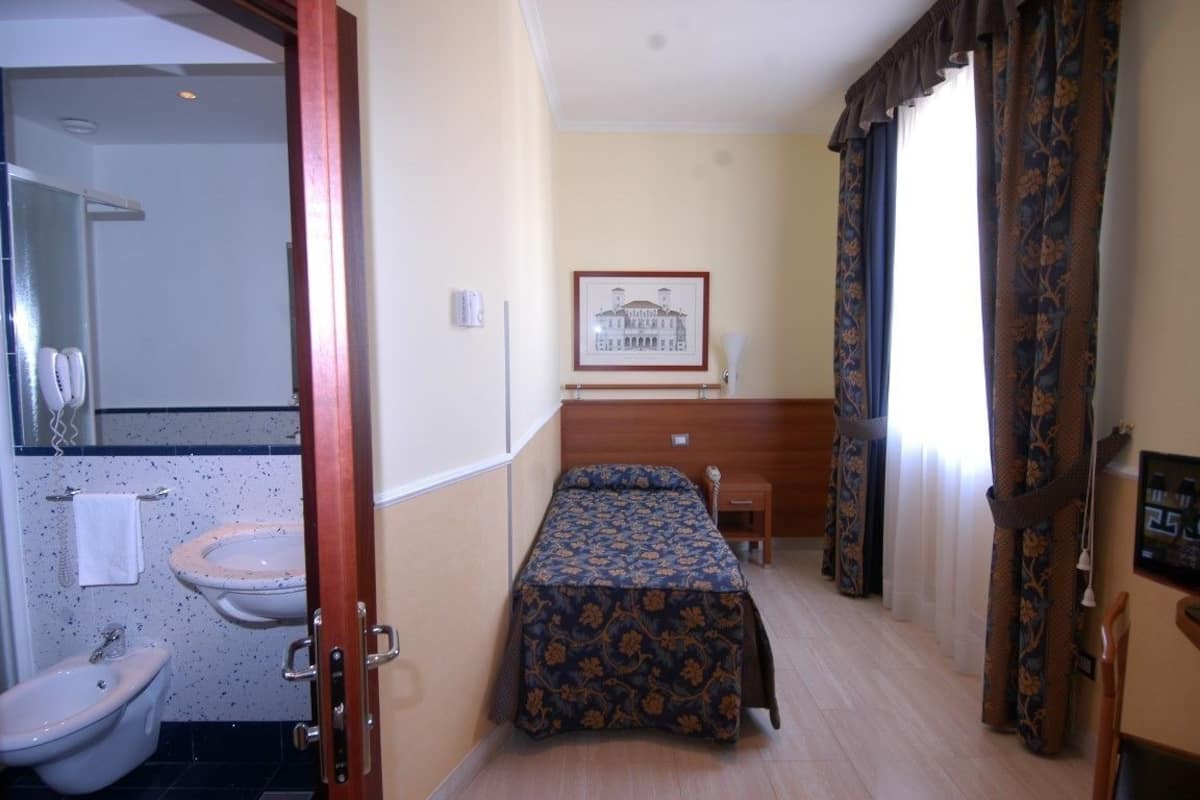 Une petite chambre d’hôtel avec un lit simple, une salle de bain comprenant un lavabo et des toilettes, et un tableau encadré au mur.