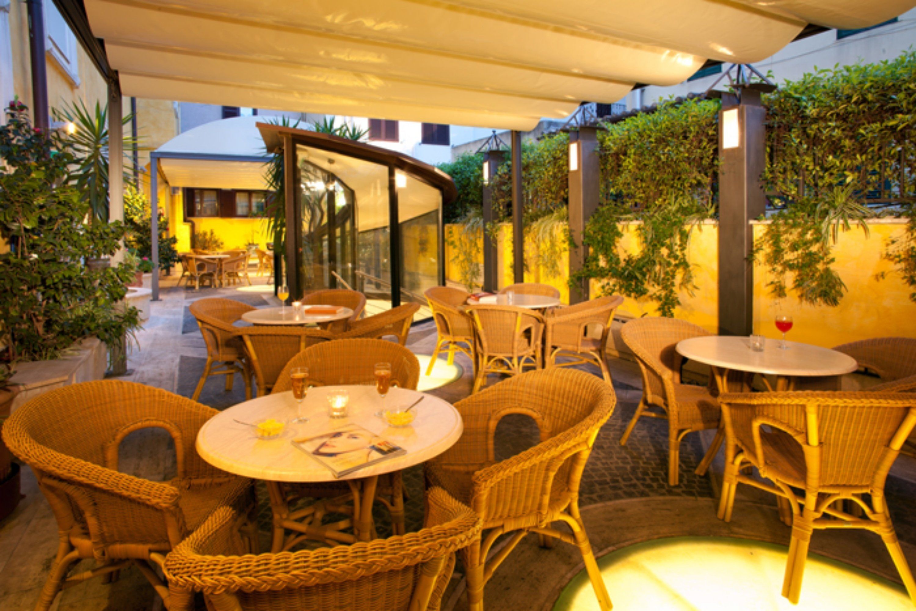 Ein Terrassenbereich im Freien mit Korbmöbeln und Tischen, umgeben von Blumentöpfen und einer Glaseinfassung.