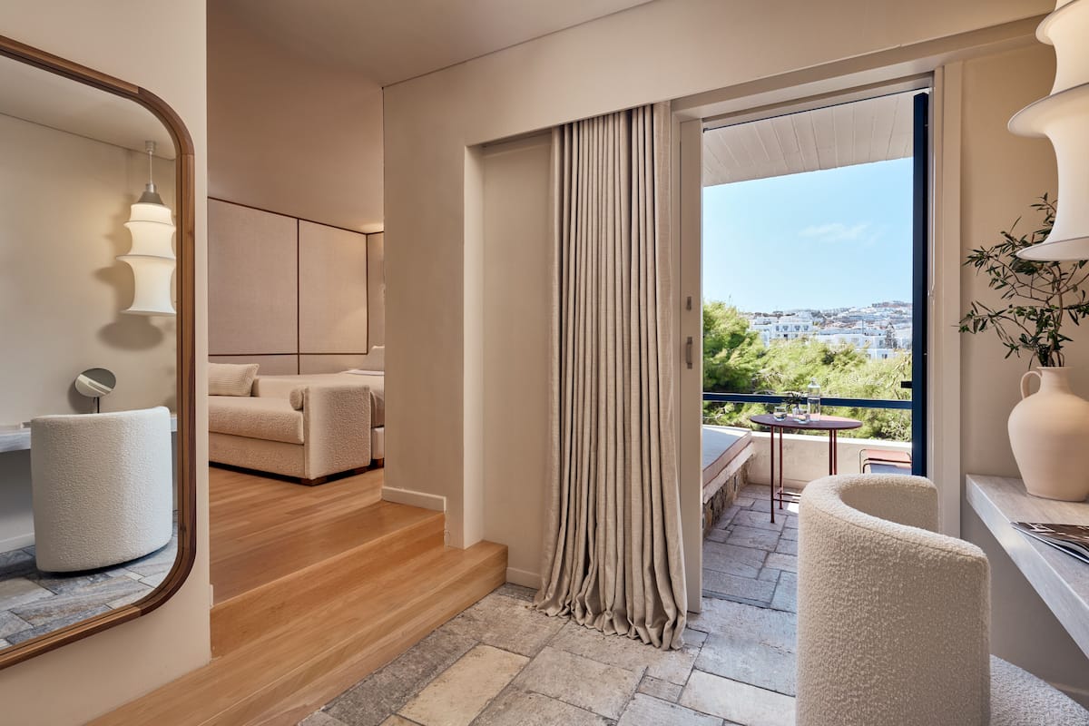 A modern hotel room with a balcony, a sofa, a chair, and a large window with curtains.