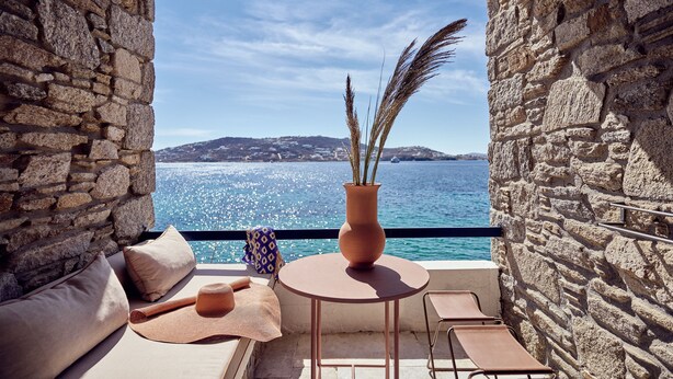 A stone-walled balcony with a round table, a vase with dried plants, and a view of the sea and distant hills.