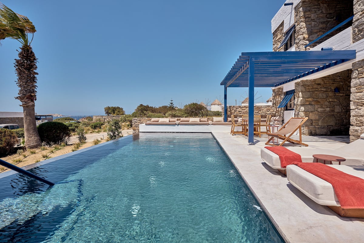 A clear blue pool with lounge chairs and a pergola, set against a stone building with a view of the sea.
