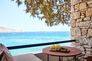 Un balcón con pared de piedra, una mesa redonda con una bandeja de frutas y bebidas, y vista al mar.