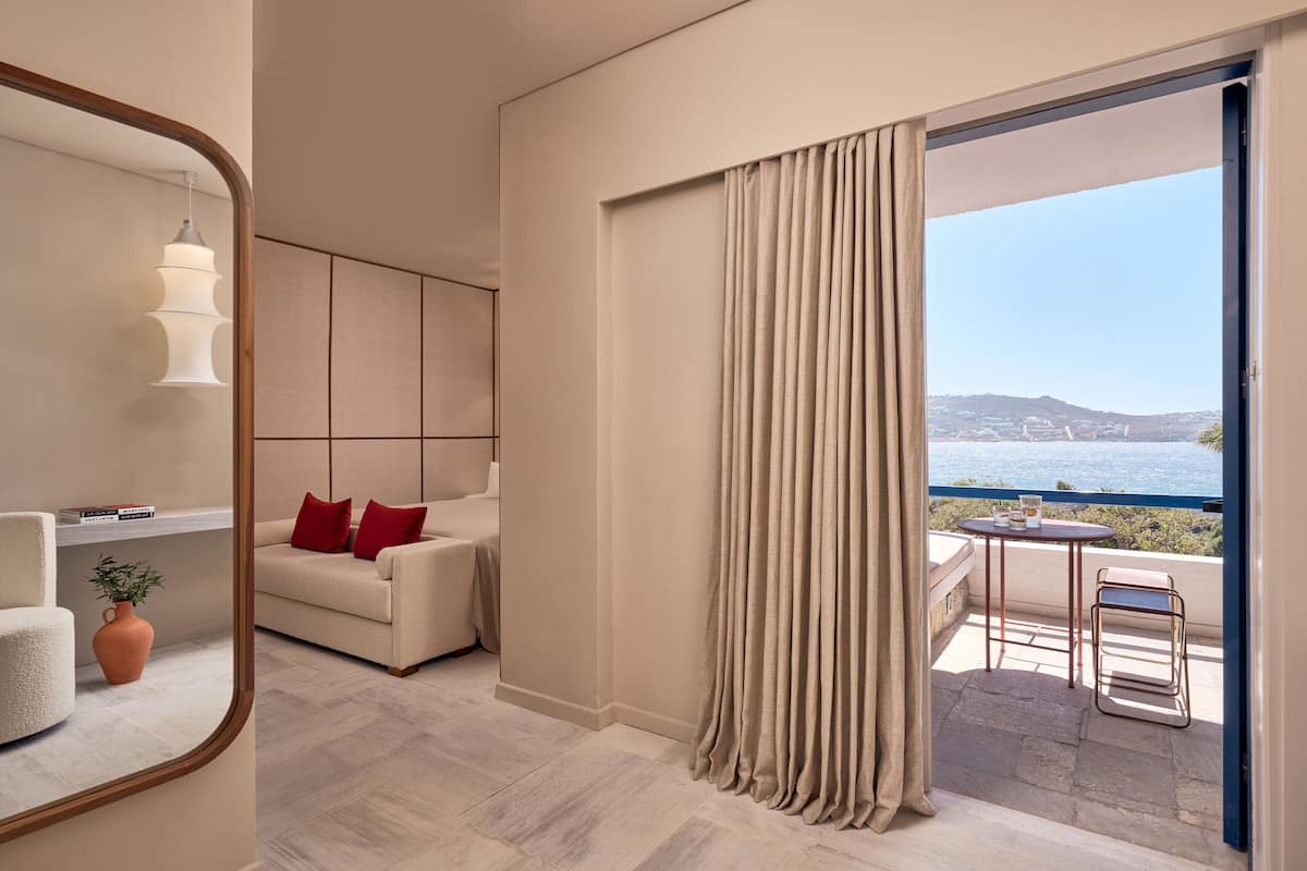 A modern hotel room with a large mirror, a beige sofa with red cushions, a small table with a chair, and a view of the sea through a balcony.