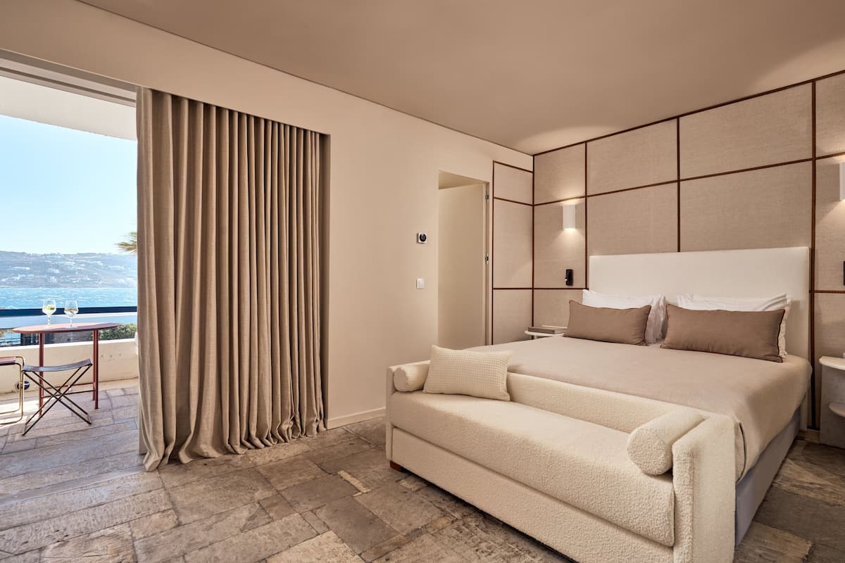 A bedroom with a large bed, a sofa, a view of the sea, and a balcony with a table and chairs.