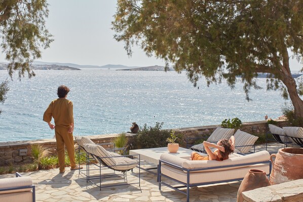 A coastal outdoor seating area with a stone floor, lounge chairs, and a view of the sea.