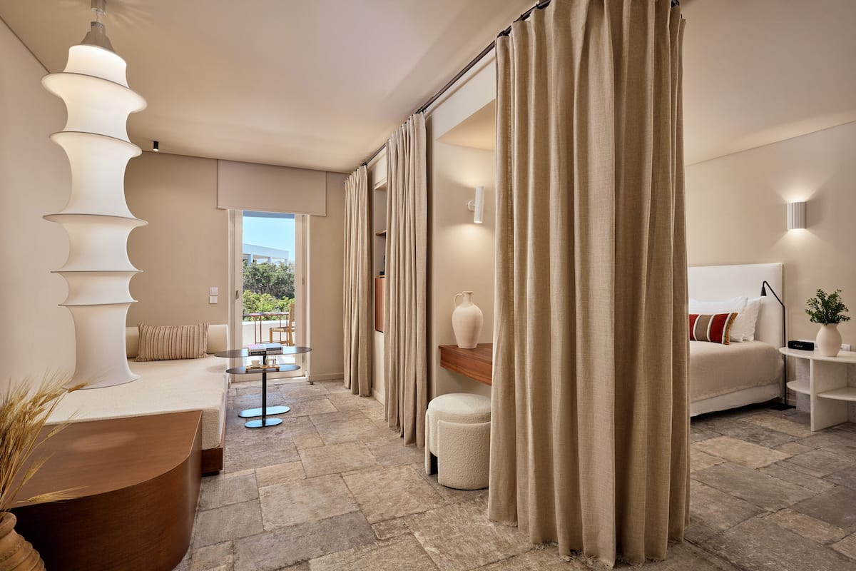 A modern hotel room with a stone floor, a sofa, a dining area, and a large window.