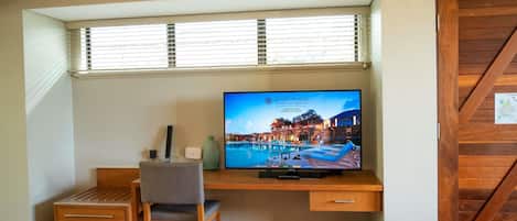 Habitación de hotel con un escritorio de madera, un televisor mostrando una imagen del hotel, una silla, una mesita y una ventana con persianas.