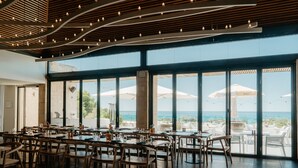 Un comedor interior con mesas y sillas, ventanas amplias con vistas al océano y un techo con luces colgantes.