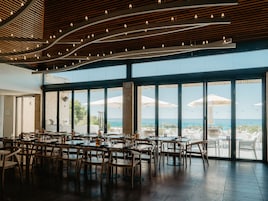 Un comedor interior con mesas y sillas, ventanas amplias con vistas al océano y un techo con luces colgantes.