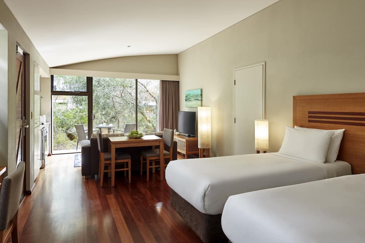 A hotel room with two beds, a desk with a chair, a television, and a window with a view of greenery.