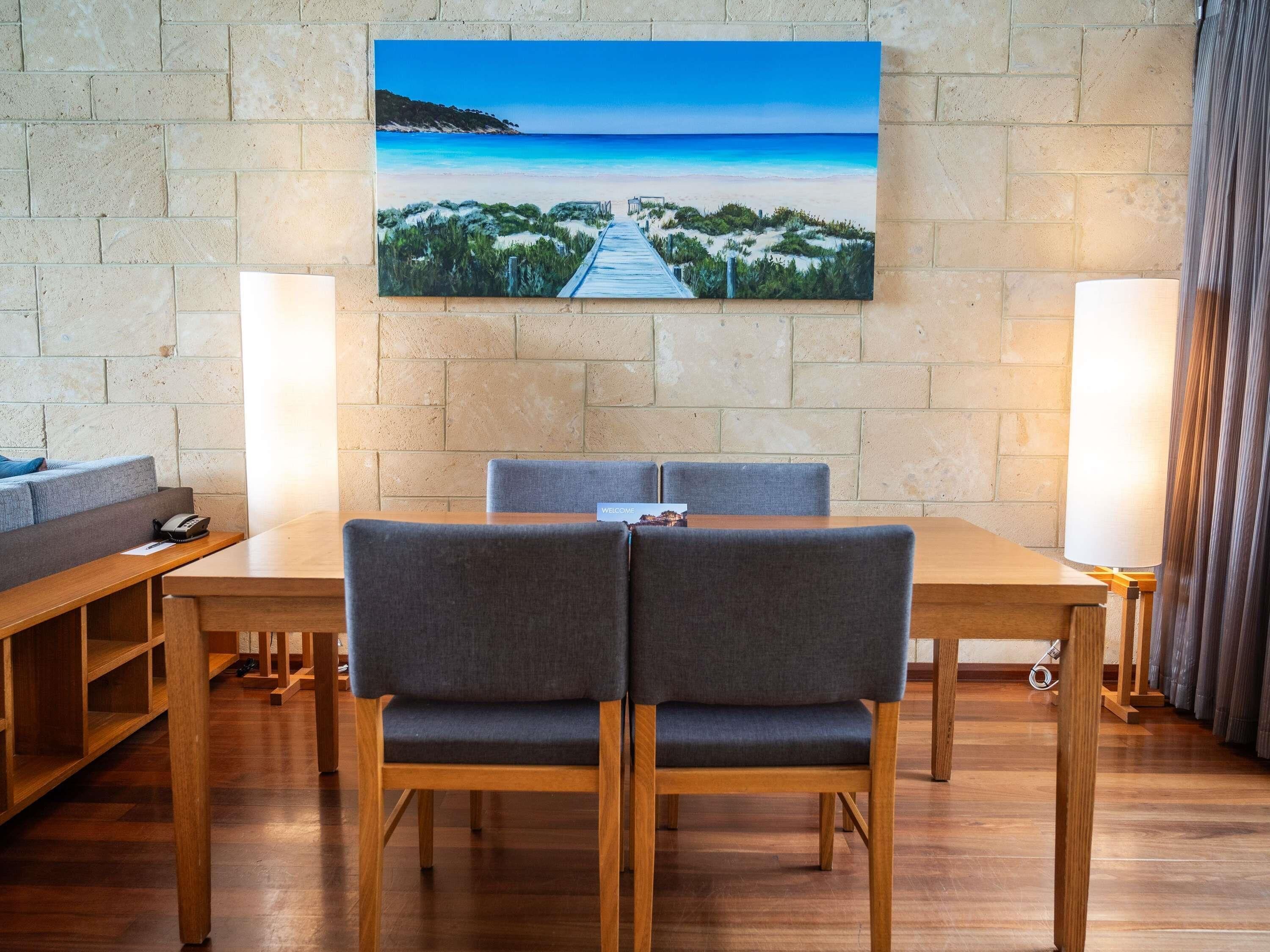 Ein Essbereich mit einem Holztisch, zwei grau gepolsterten Stühlen, einer Bank und einem Bild einer Strandansicht an der Wand.