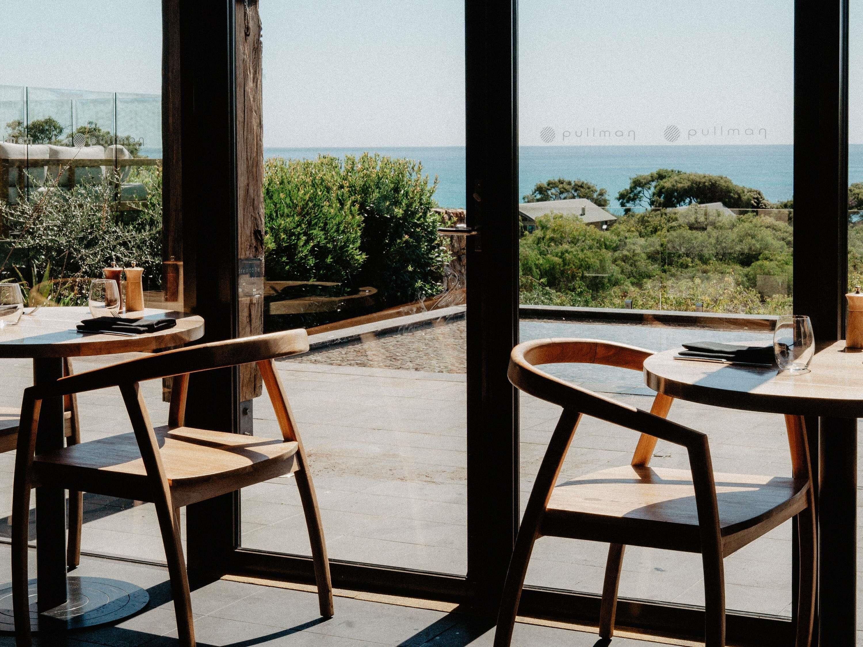 Un espace de restauration en plein air, avec des chaises et des tables en bois, offrant une vue panoramique sur la mer et la végétation.