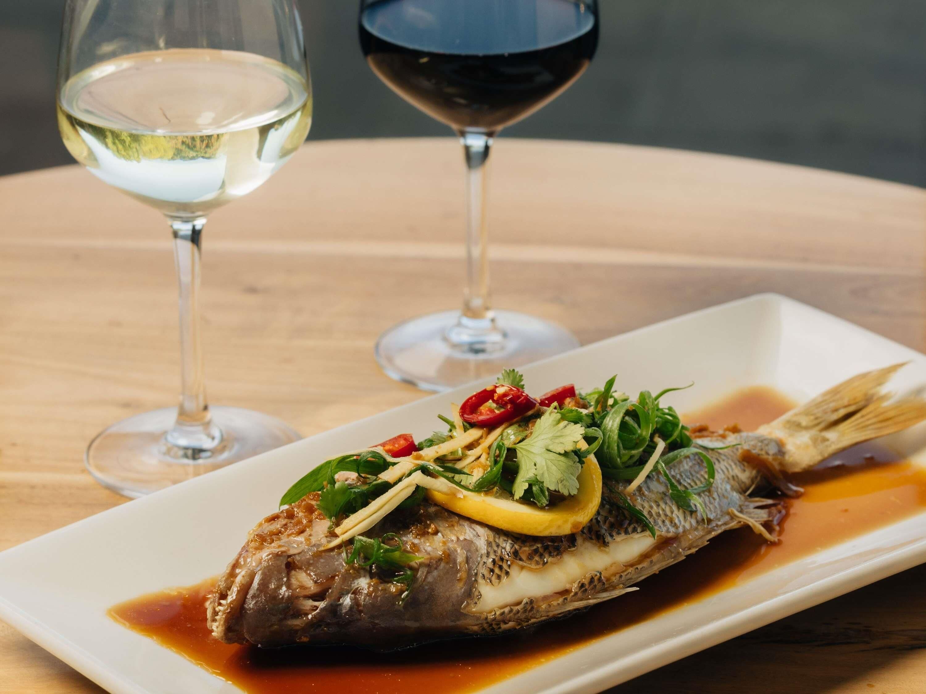 En vit, rektangulär tallrik med en tillagad fisk, garnerad med örter och citronskivor, tillsammans med två glas vin på ett träbord.