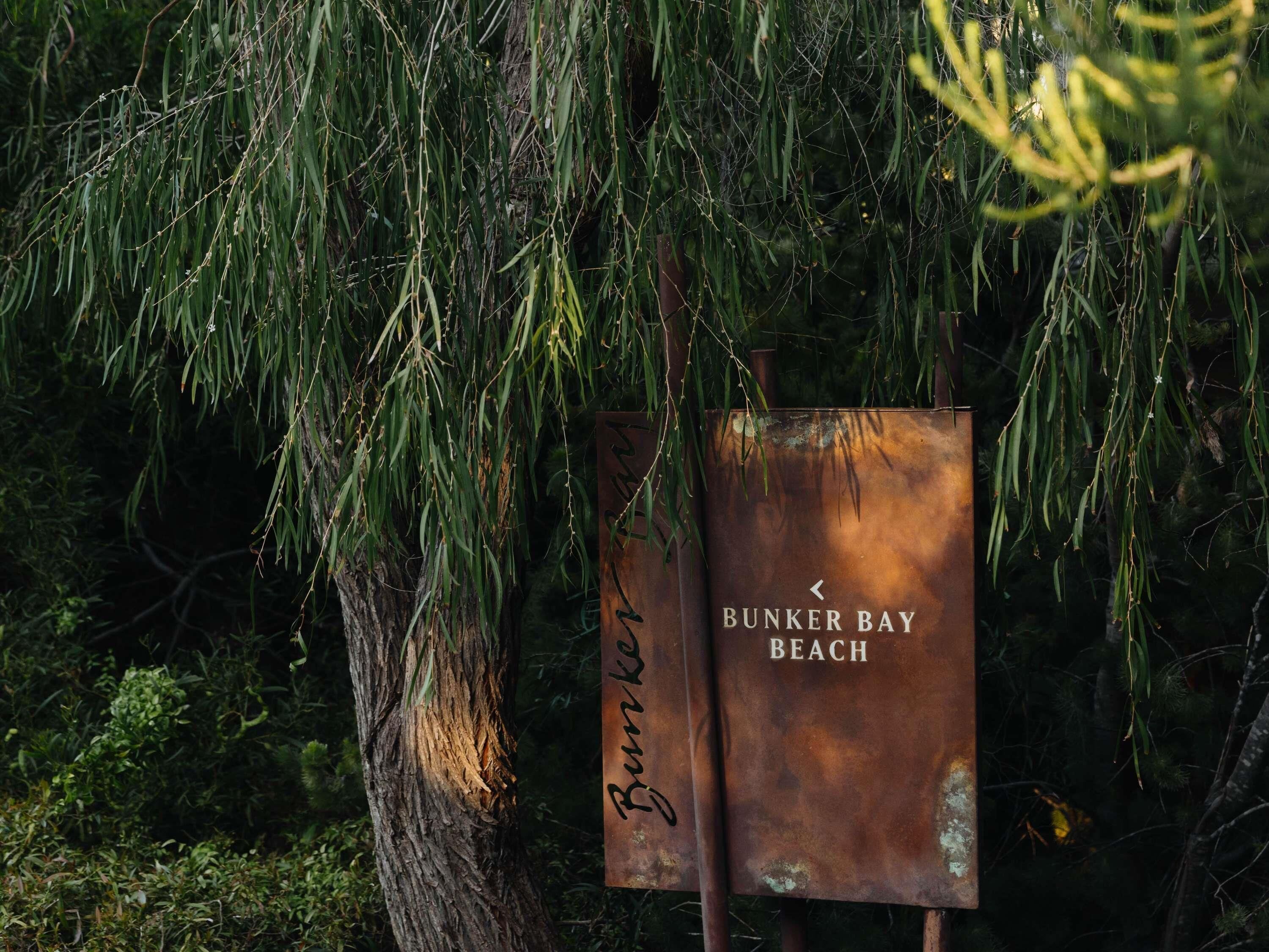 A rusted metal sign with 'Bunker Bay Beach' written on it, hanging amidst dense foliage.