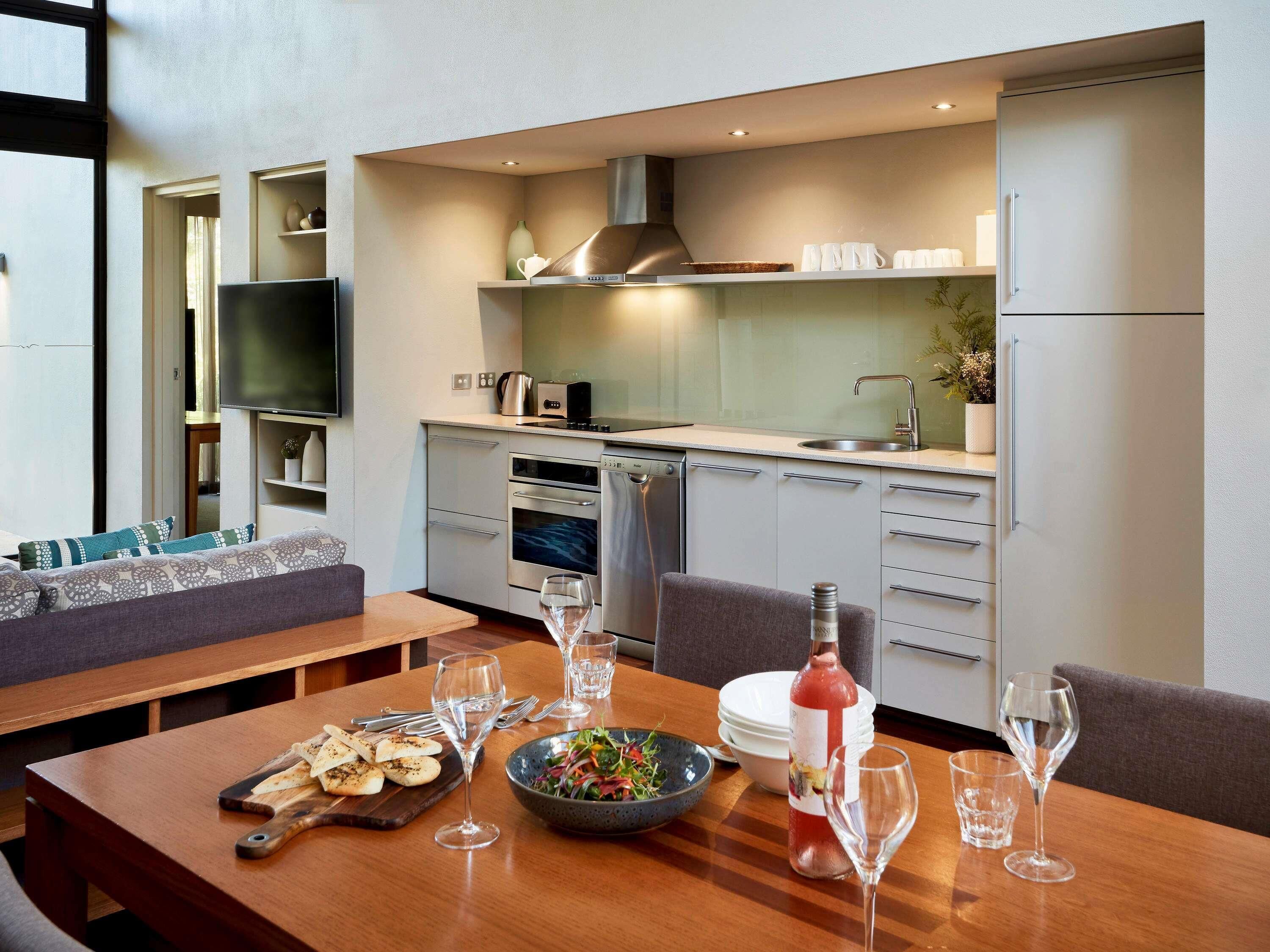 Una cocina moderna con electrodomésticos integrados, un comedor con mesa de madera lista para una comida, y una sala de estar con sofá y televisor.