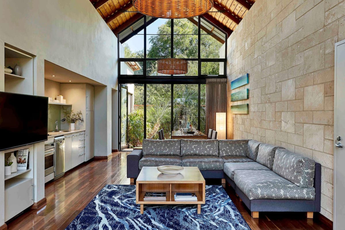 A modern living room with a large sectional sofa, a flat-screen TV, a dining area with a table and chairs, and a kitchen area with cabinets and a TV.