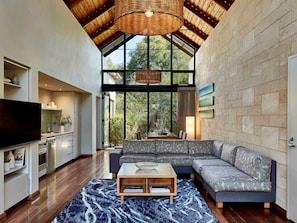 A modern living room with a large sectional sofa, a flat-screen TV, a dining area with a table and chairs, and a kitchen area with cabinets and a TV.