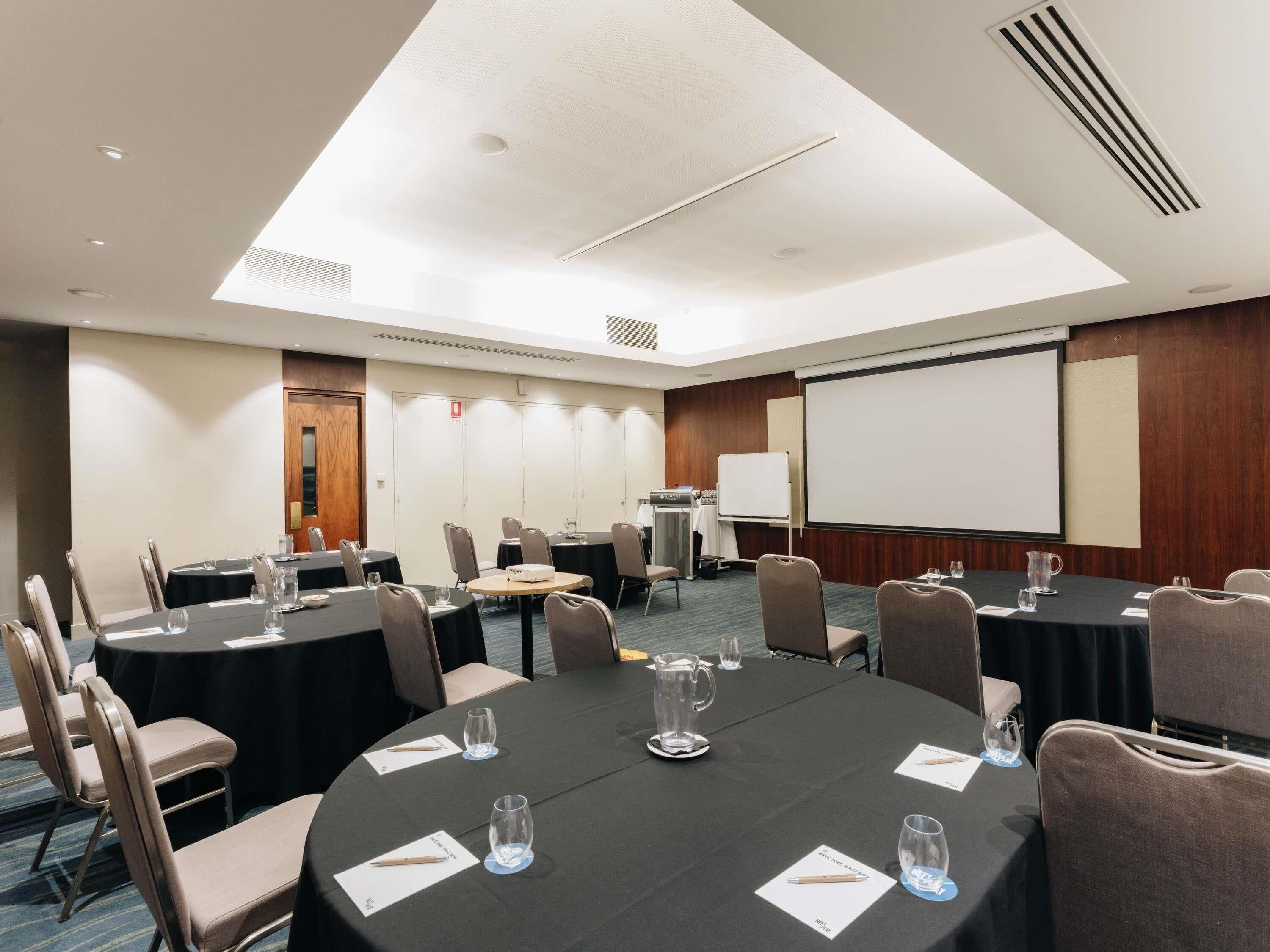 Une salle de conférence équipée de tables rondes, de chaises, d’un écran de projection et d’un tableau blanc.