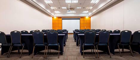 Un salón de conferencias con filas de sillas, una pantalla para proyector y suelo alfombrado.
