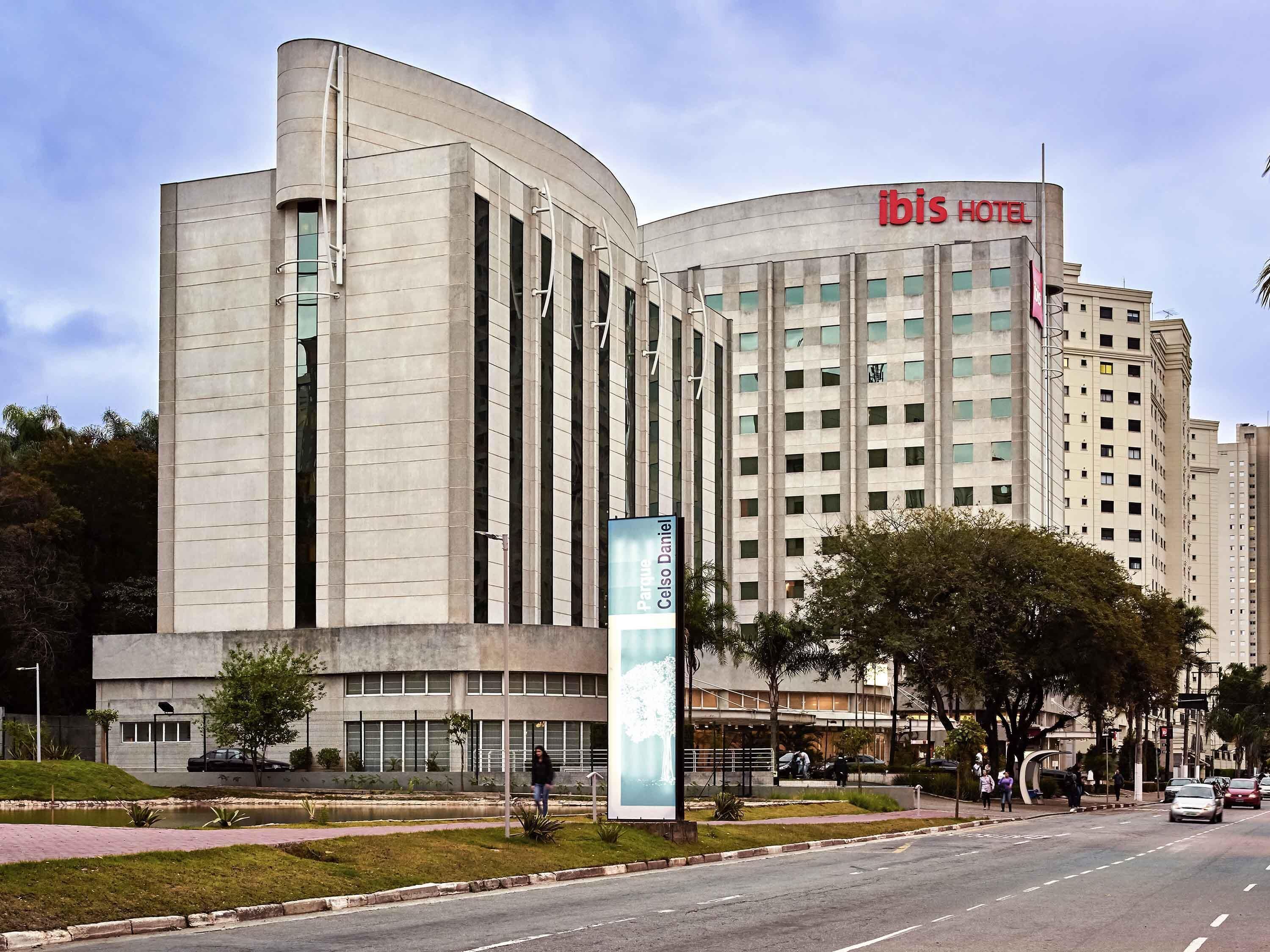 Un hotel moderno con fachada curva, un llamativo letrero 'ibis HOTEL' y una zona ajardinada con un sendero y árboles.