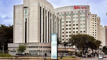Un hotel moderno con fachada curva, un llamativo letrero 'ibis HOTEL' y una zona ajardinada con un sendero y árboles.