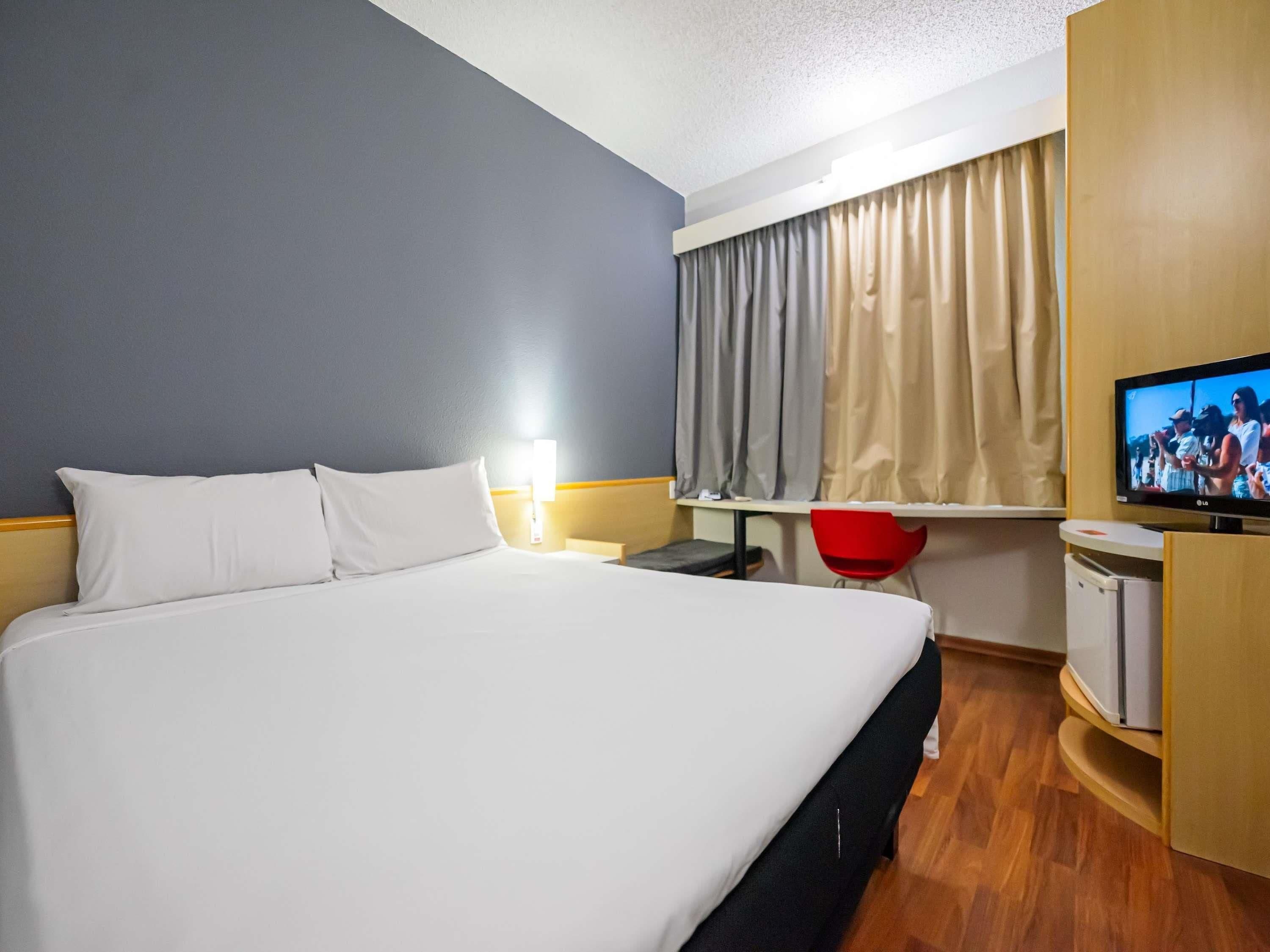 A hotel room with a large bed, a desk with a chair, a television, and a window with curtains.