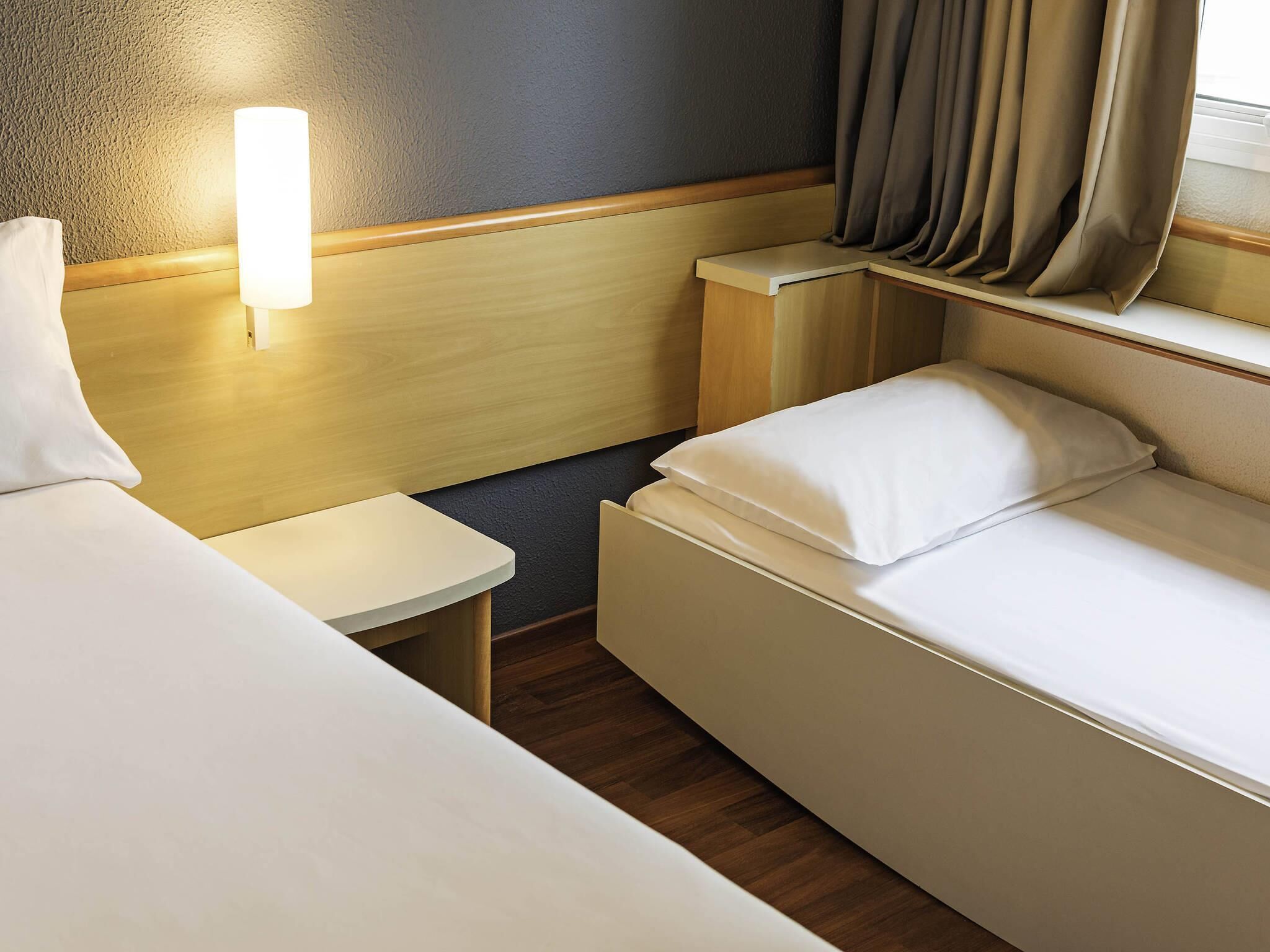 A hotel room with two beds, a bedside table, a lamp, and a window with curtains.