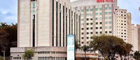 Edificio moderno de hotel con fachada curva, un llamativo letrero 'ibis HOTEL' y un área ajardinada con fuente de agua y árboles.
