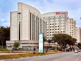 A modern hotel building with a curved facade, a prominent 'ibis HOTEL' sign, and a landscaped area with a water feature and trees.