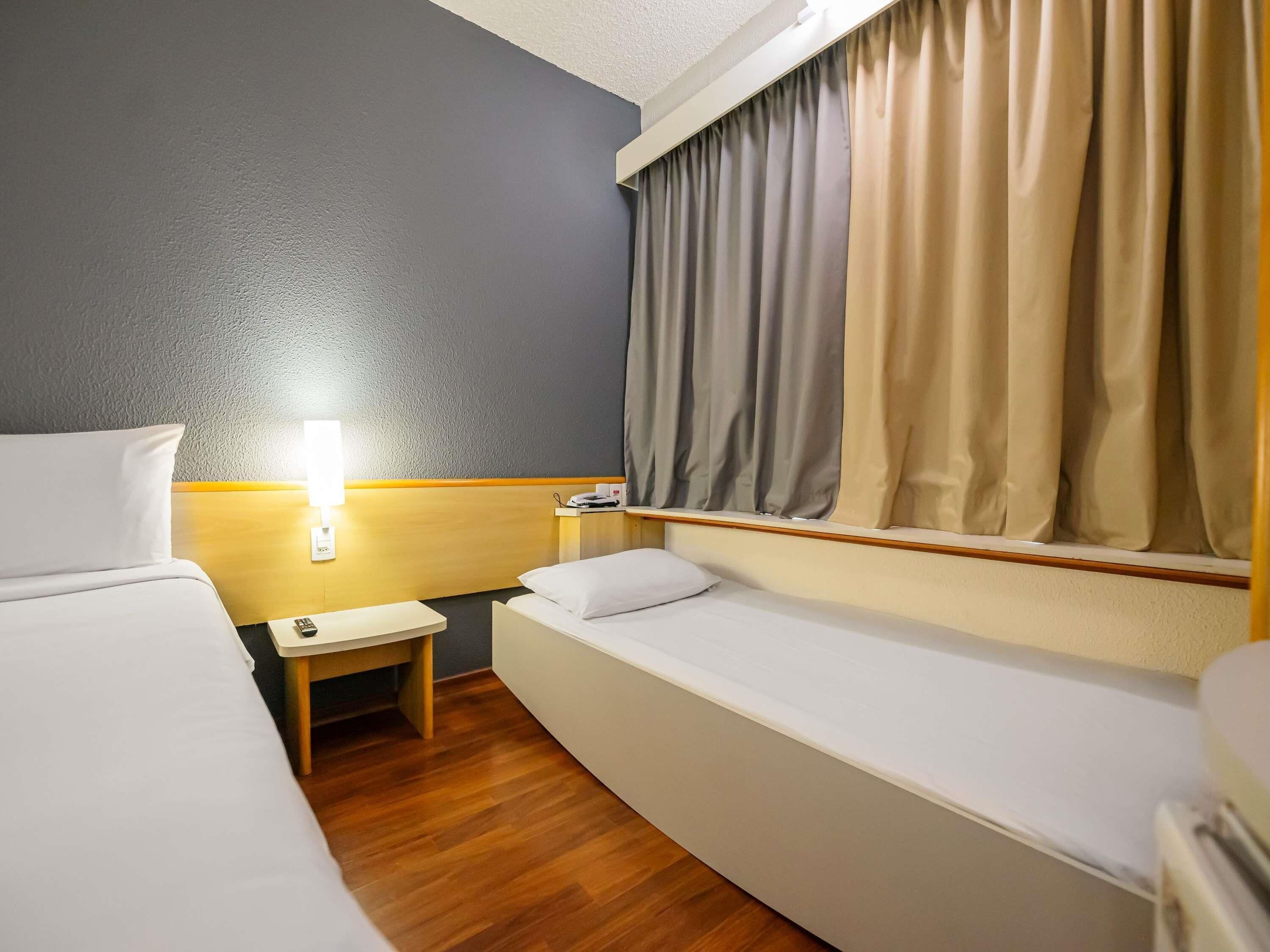 A hotel room with two beds, a small bedside table, a lamp, and a window with curtains.