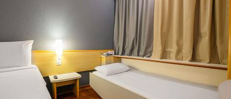 A hotel room with two beds, a small bedside table, a lamp, and a window with curtains.