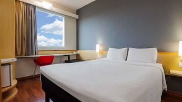 Ett hotellrum med en säng, ett skrivbord, en stol och ett fönster med gardiner.