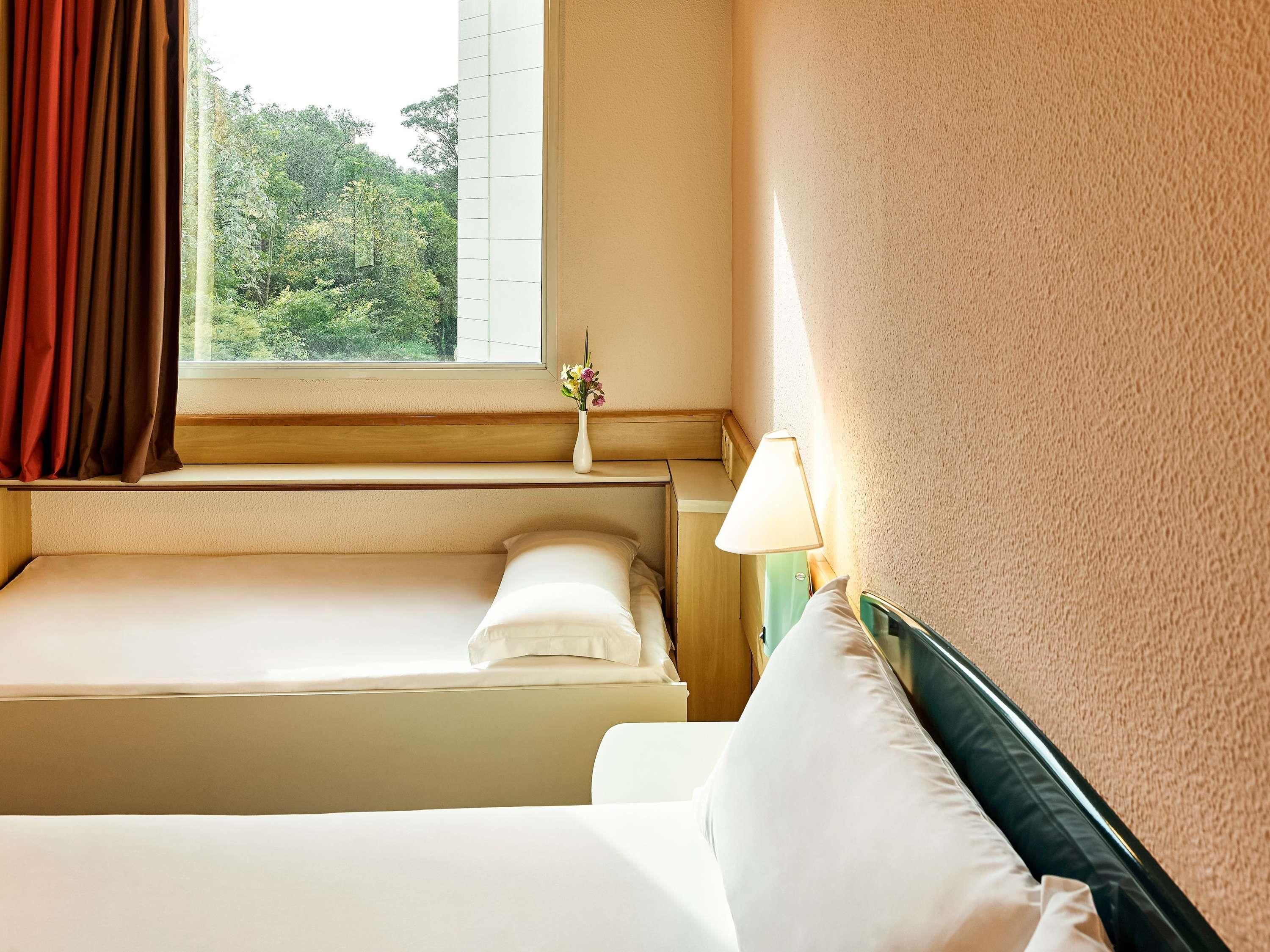 A hotel room with a bed, a bedside table with a lamp, and a window with curtains.