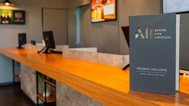 Ein Empfangstresen mit einem Schild, auf dem 'PRIORITY WELCOME' und 'ACCOR LIVE LIMITLESS' steht, in einem modernen Innenraum mit Pendelleuchten.