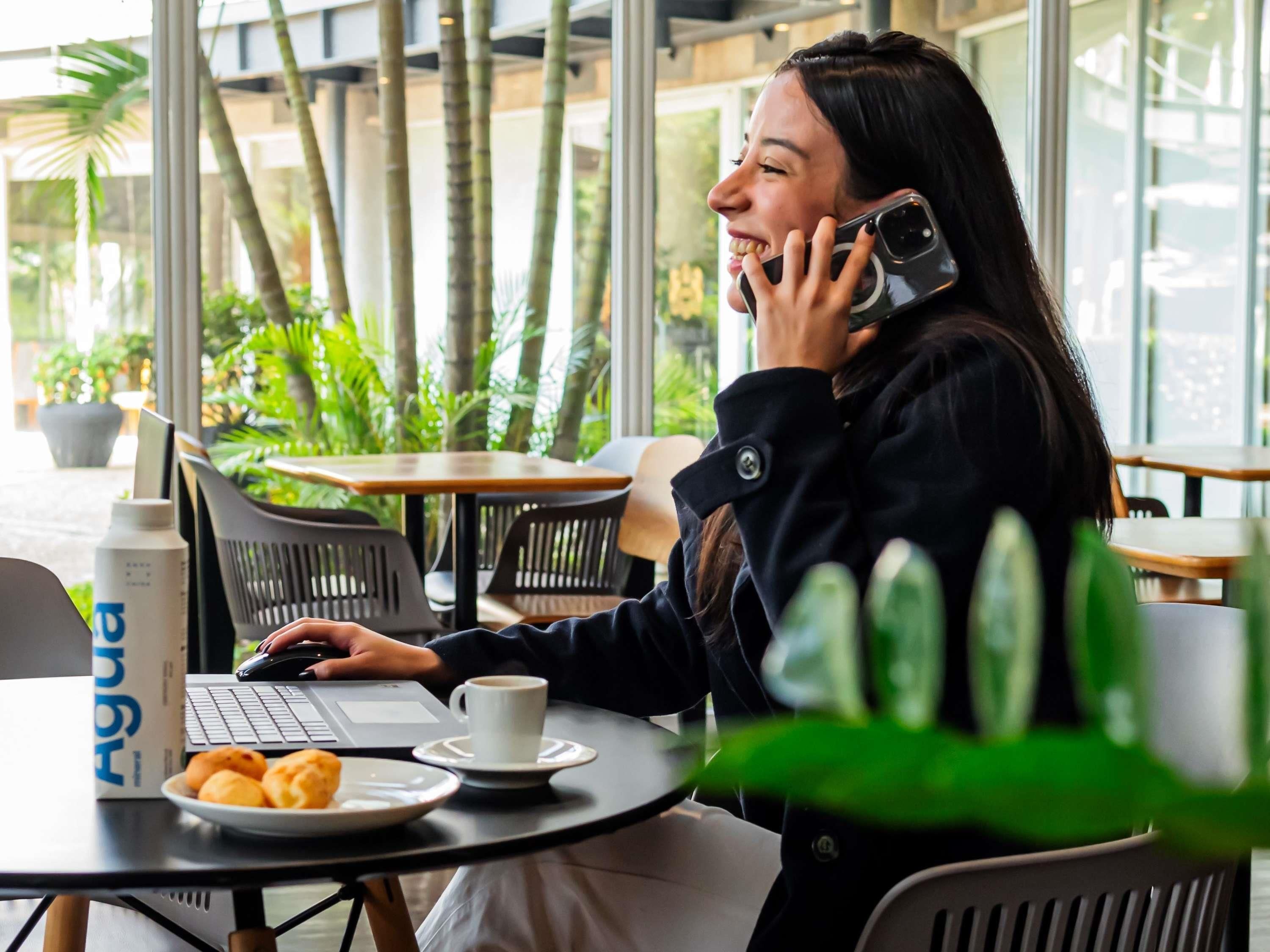 A person using a laptop at a table with a cup and pastries, in a cafe with indoor plants and a glass window.