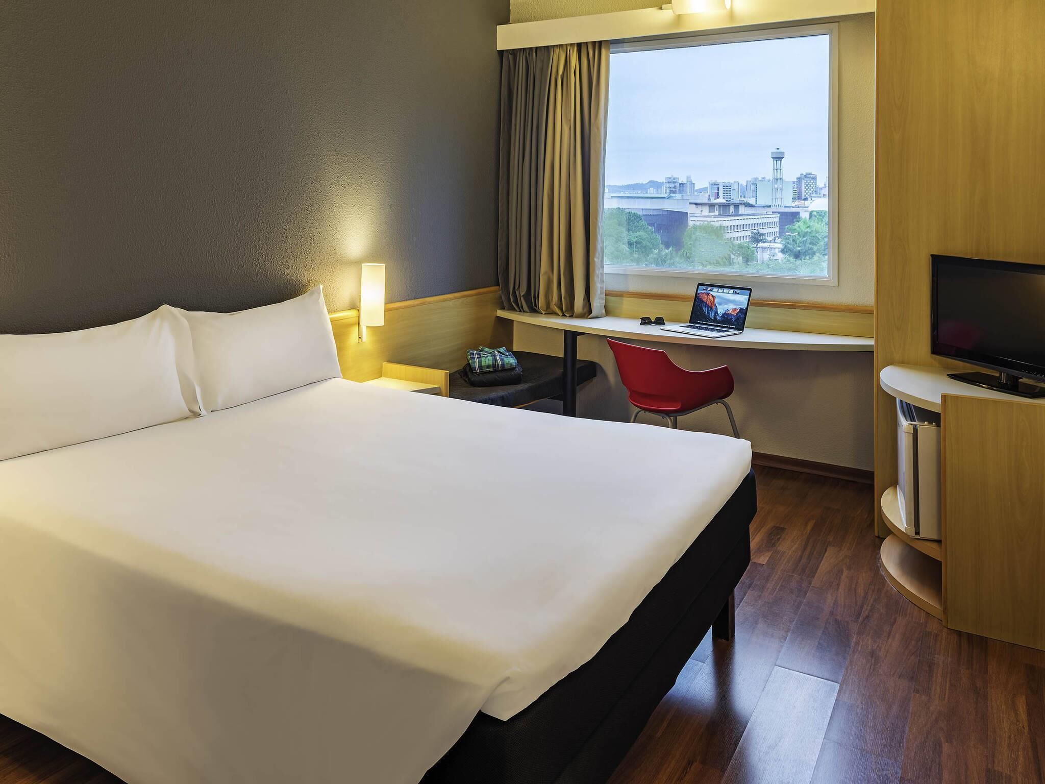 A hotel room with a large bed, a desk with a laptop, a TV, and a city view.