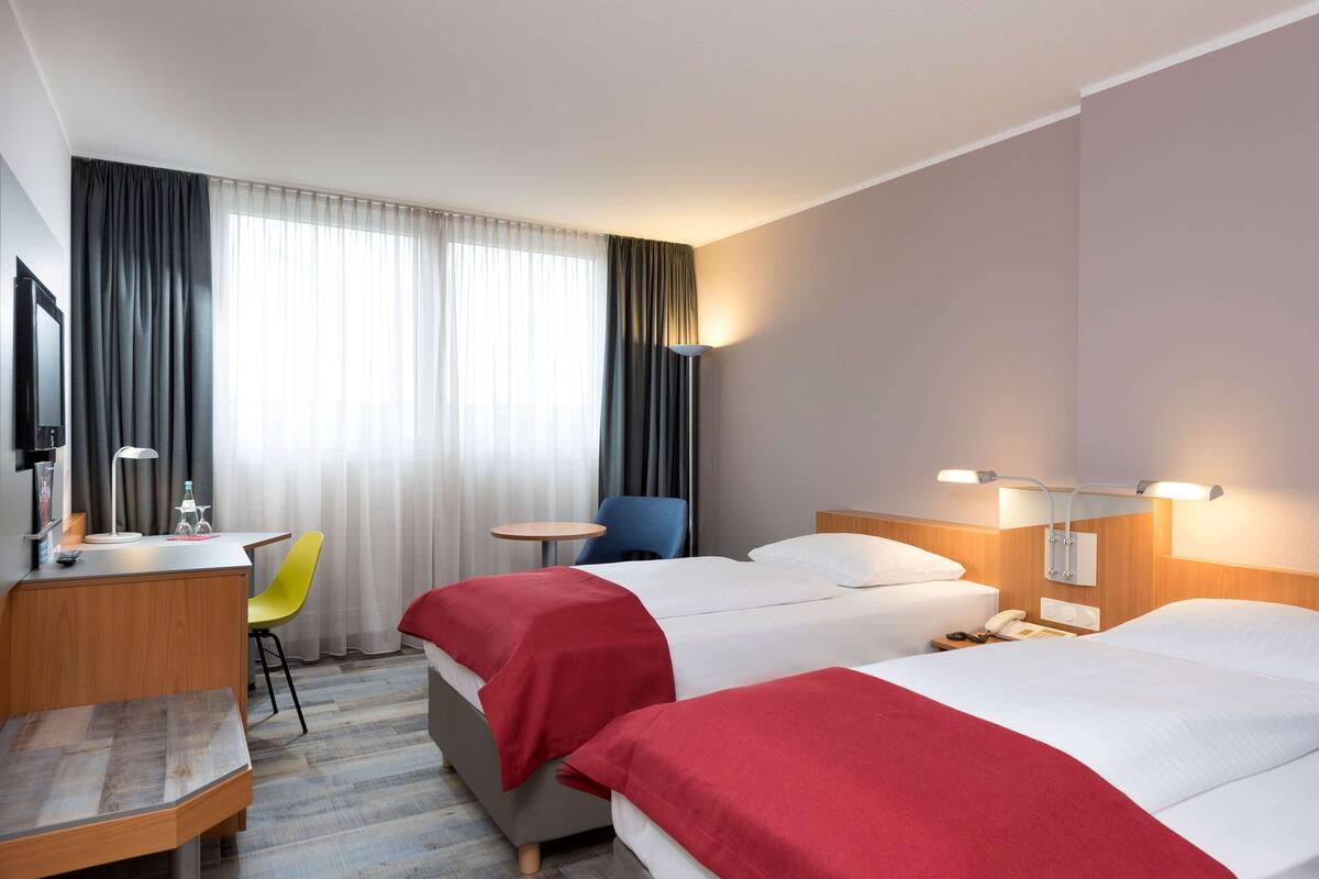 A hotel room with two beds, a desk, a chair, a small table, and a TV.