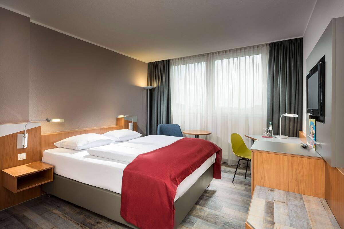 A hotel room with a bed, bedside table, chair, desk, and a TV.