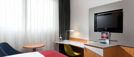 A hotel room with a bed, a desk with a chair, a television, and a window with curtains.