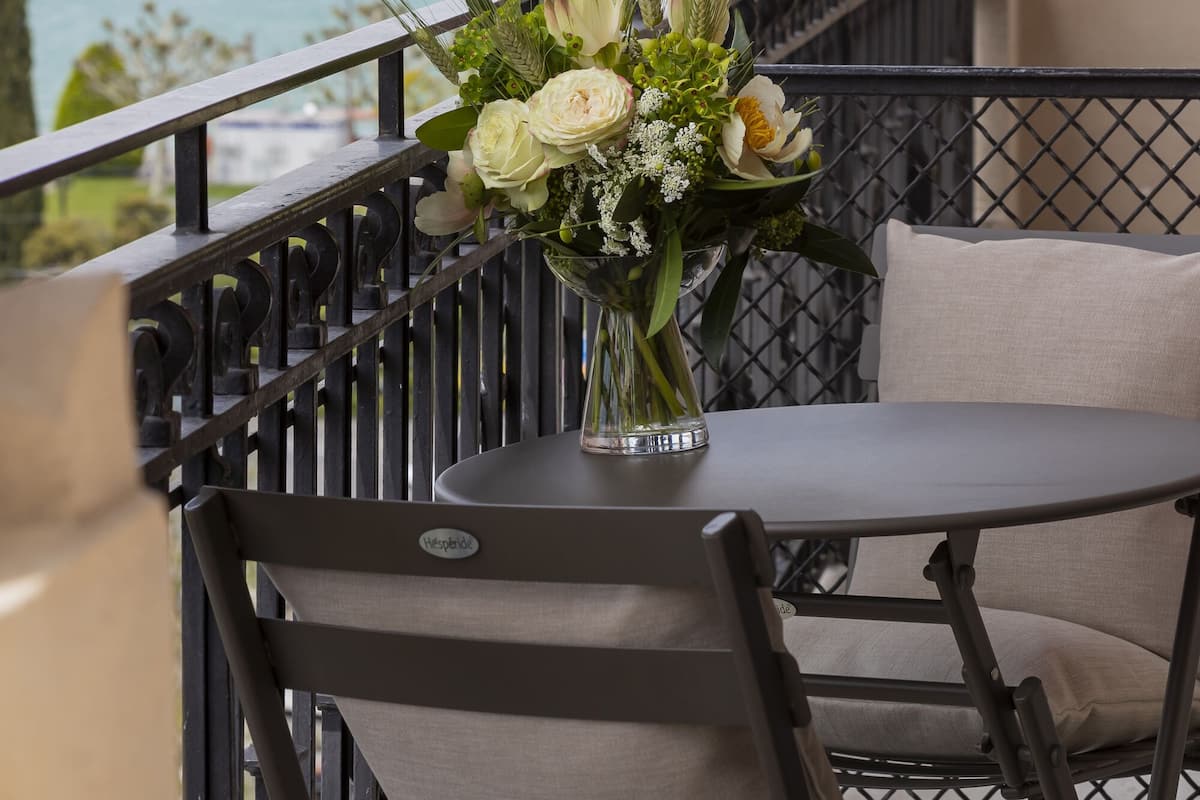 A balcony with a table and chairs, a vase of flowers, and a view of the sea and buildings.