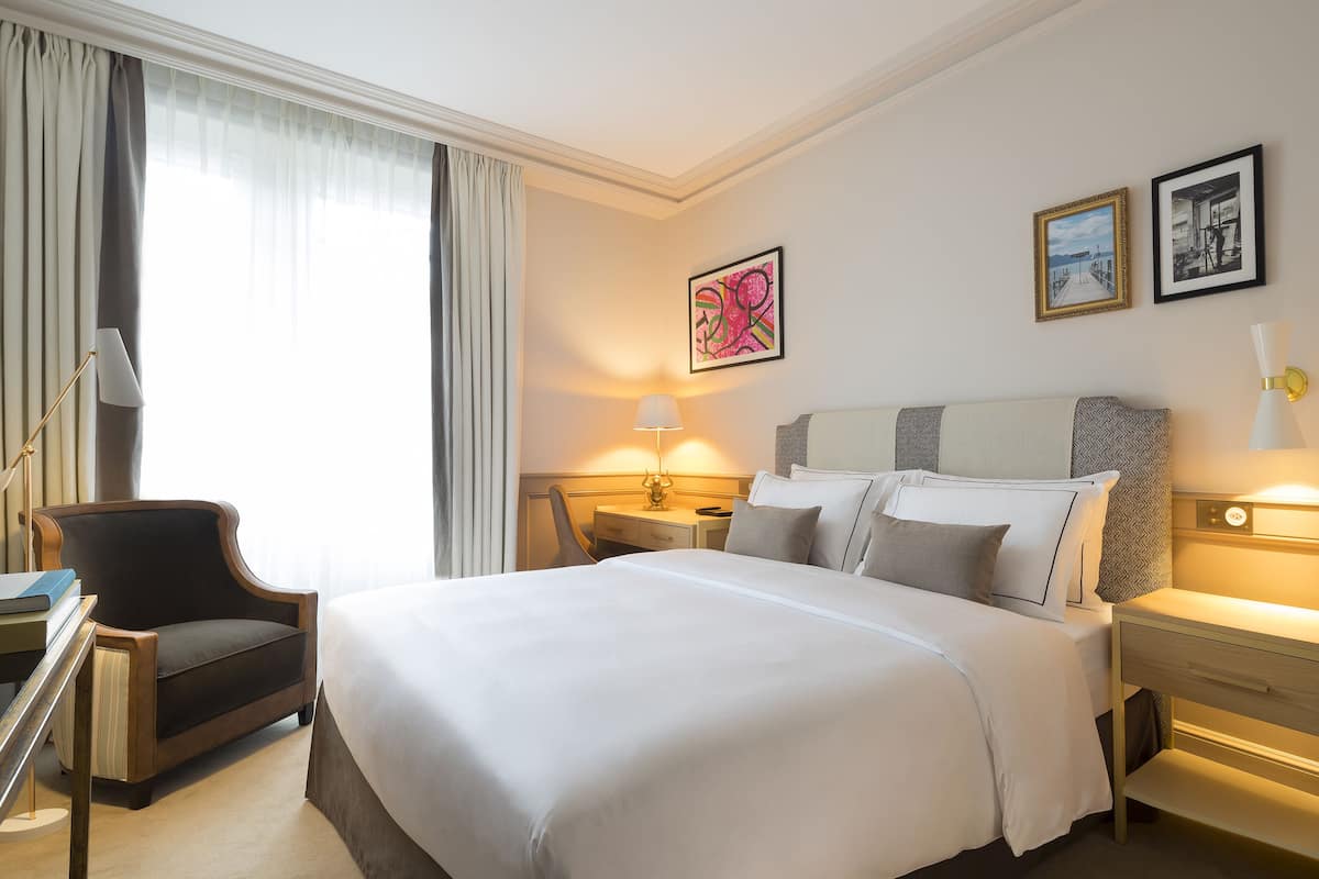 A hotel room with a large bed, a desk with a lamp, a chair, and a nightstand with a lamp.