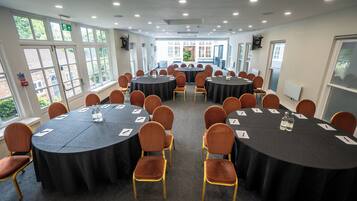 Un salón de conferencias con varias mesas redondas, cada una rodeada de sillas, y ventanas amplias que ofrecen luz natural.
