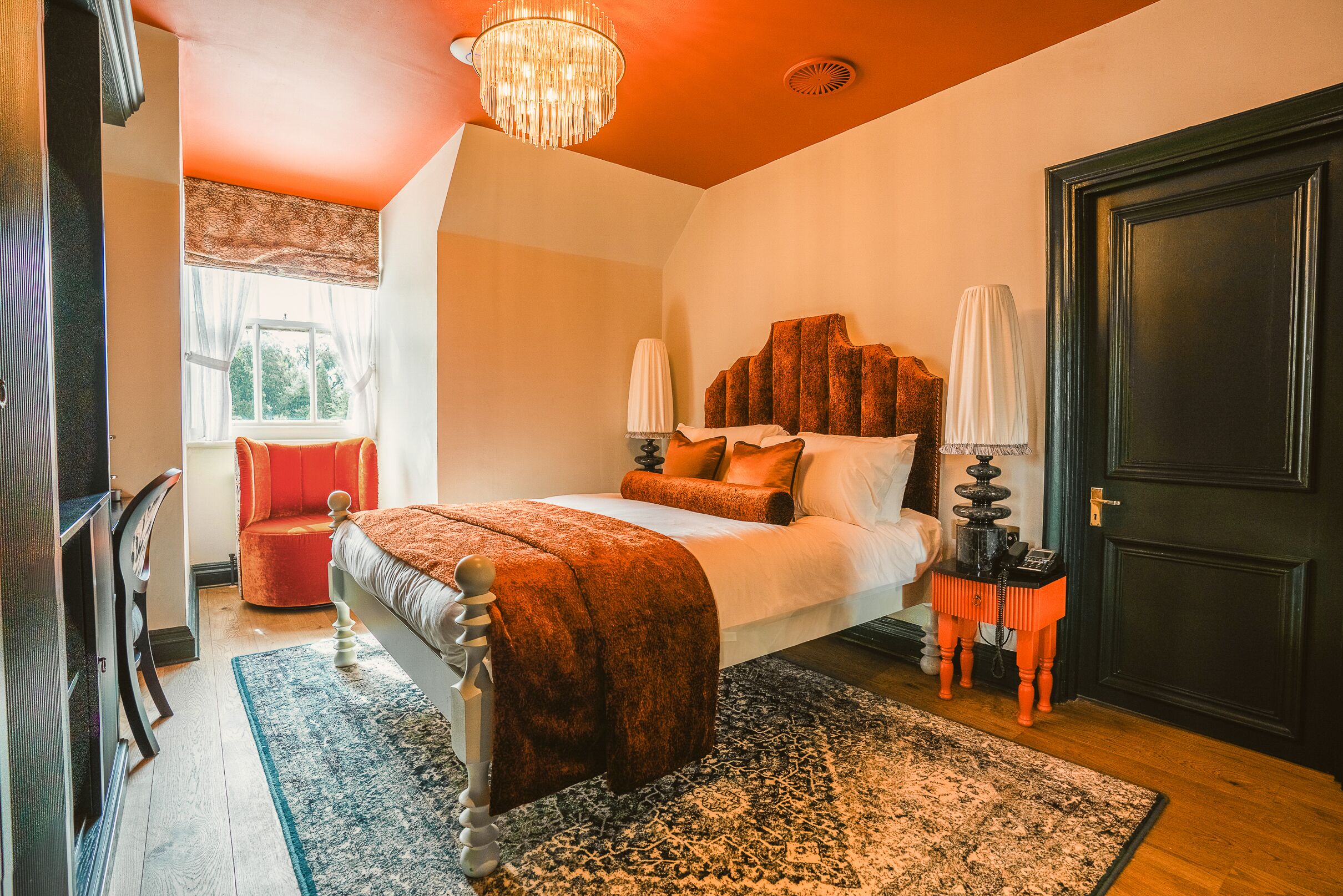 A bedroom with a bed, a chandelier, a window with curtains, a chair, and a door.