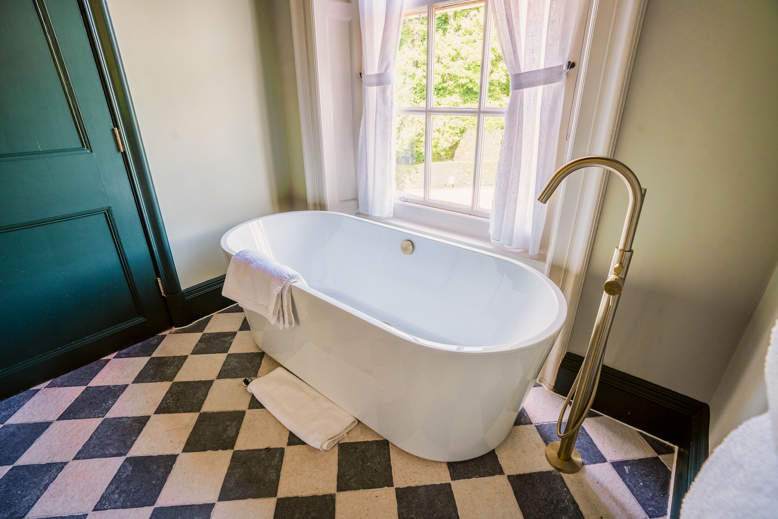 A bathroom with a freestanding bathtub, a window with curtains, and a checkered floor.
